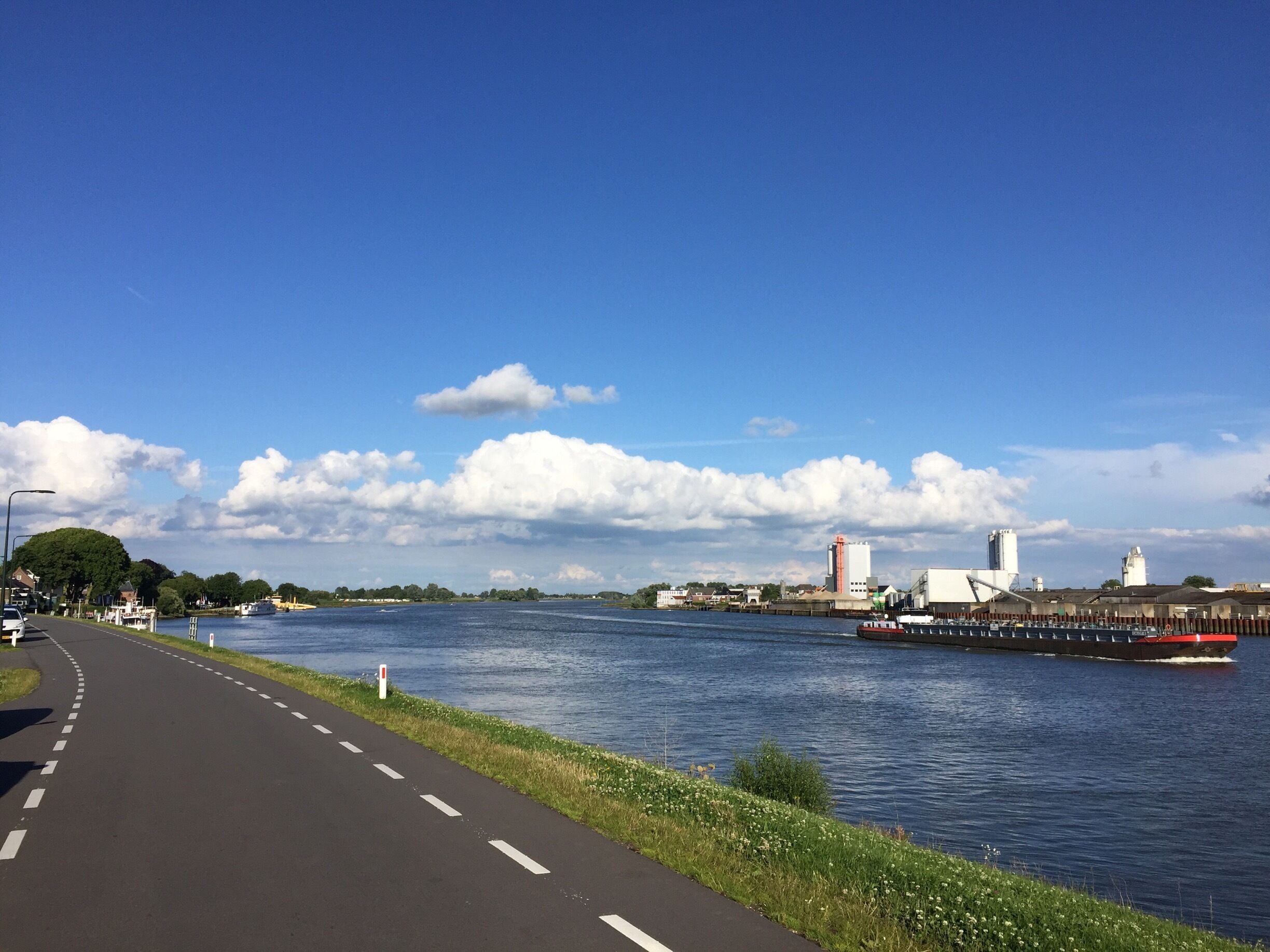 A river with a paved walkway alongside, a bridge, and industrial buildings in the background.
