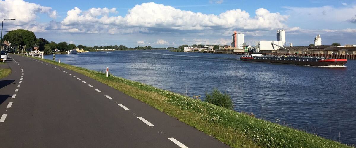 A river with a paved walkway alongside, a bridge, and industrial buildings in the background.