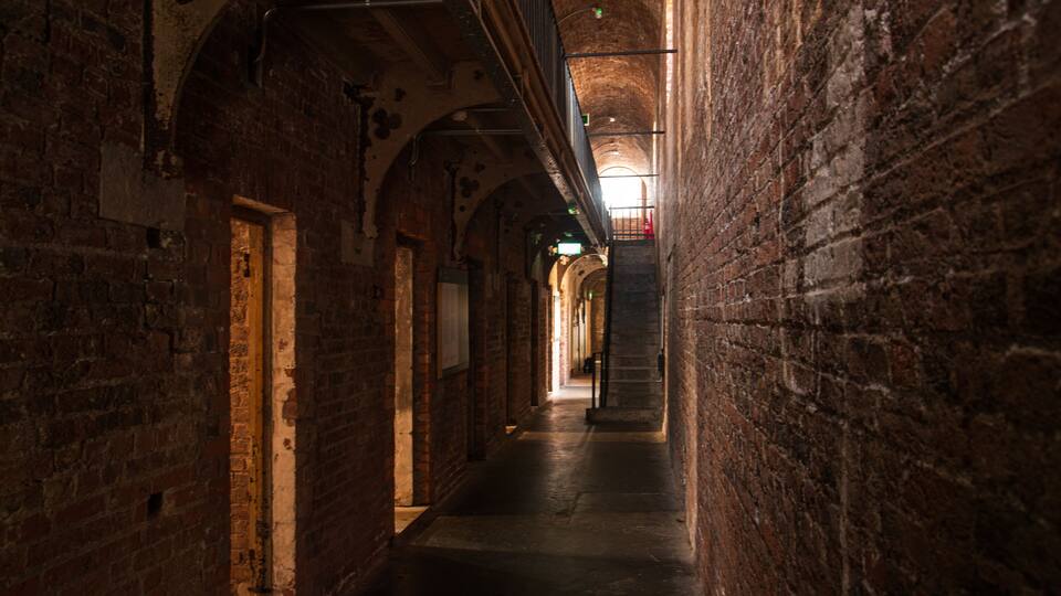 A narrow, dimly lit brick corridor with arched ceilings and a visible exit sign.