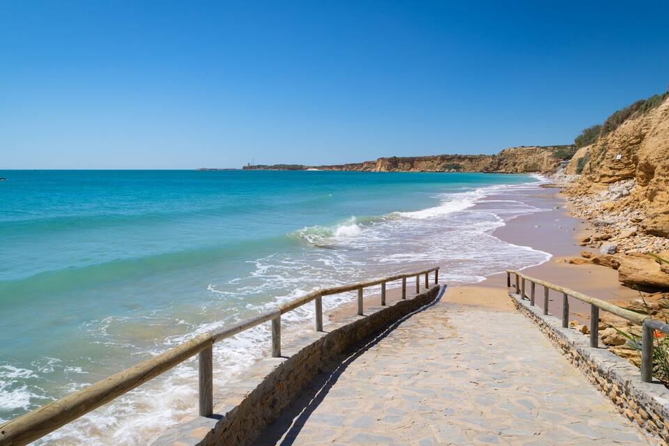 Een kustpad dat naar een strand leidt, met helder turquoise water en een rotsachtige klif.