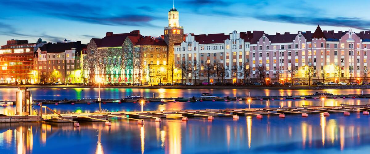A waterfront cityscape at dusk with illuminated buildings reflecting on the water.