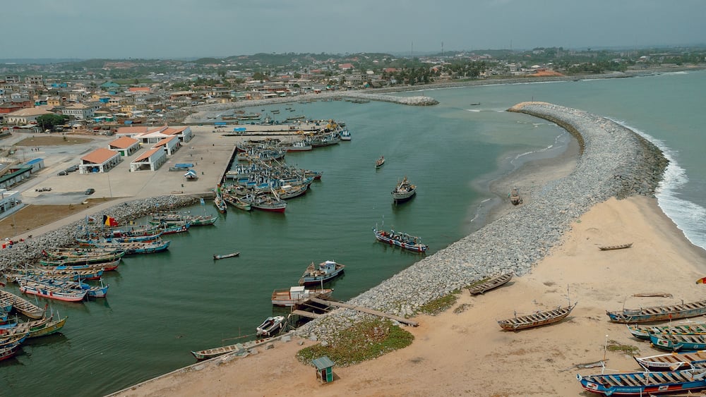 Aerial view of fishing boats clustered in the harbor contrasting with the sandy beach and stone breakwater under a cloudy sky, Elmina, Central Region, Ghana.