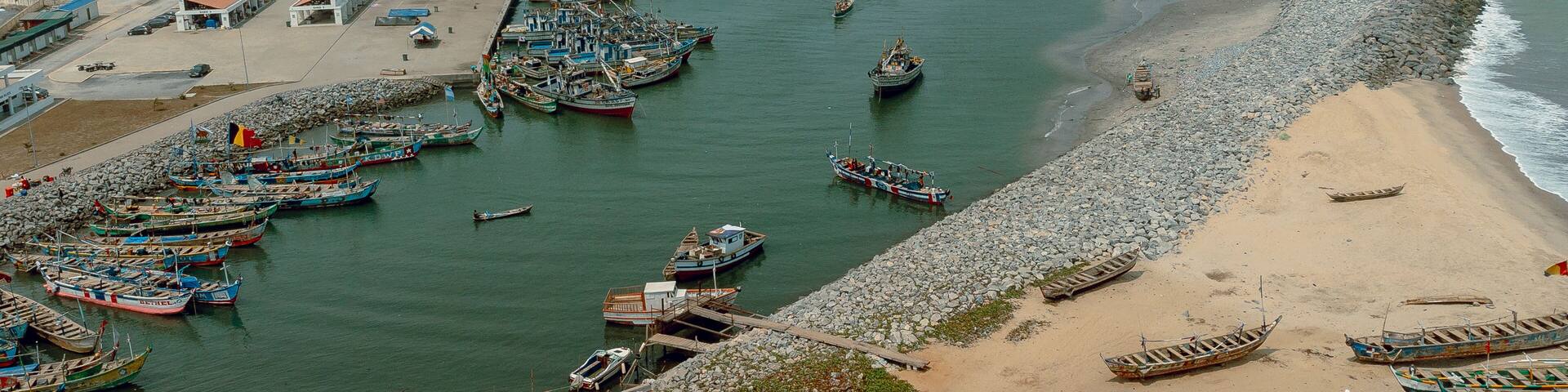 Aerial view of fishing boats clustered in the harbor contrasting with the sandy beach and stone breakwater under a cloudy sky, Elmina, Central Region, Ghana.
