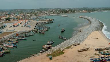 Aerial view of fishing boats clustered in the harbor contrasting with the sandy beach and stone breakwater under a cloudy sky, Elmina, Central Region, Ghana.