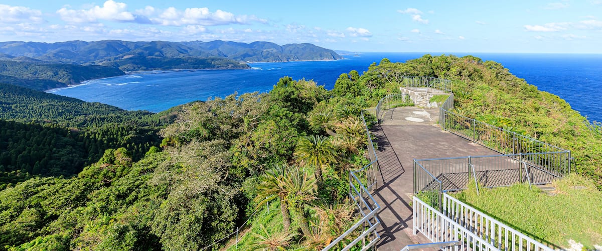 A coastal landscape with a clear view of the ocean, a forested hillside, and a paved viewing platform with railings.