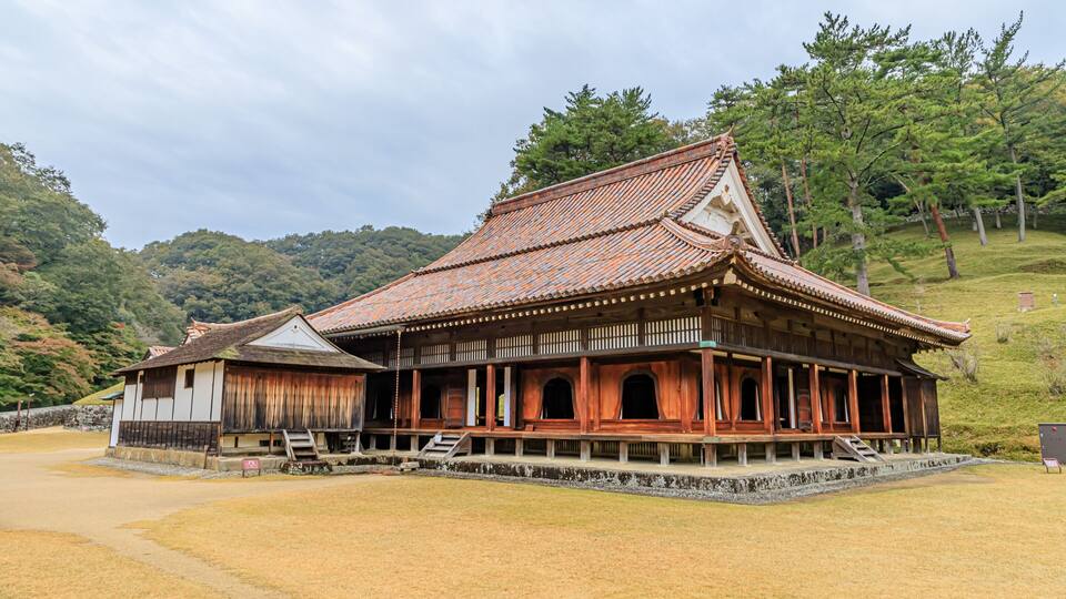 A traditional Japanese building with a tiled roof, wooden structure, and a surrounding grassy area.