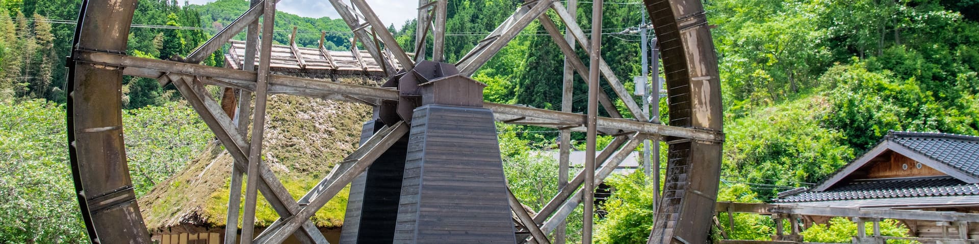 A large water wheel structure with a metal framework and wooden paddles, set against a backdrop of lush green hills and a clear blue sky.