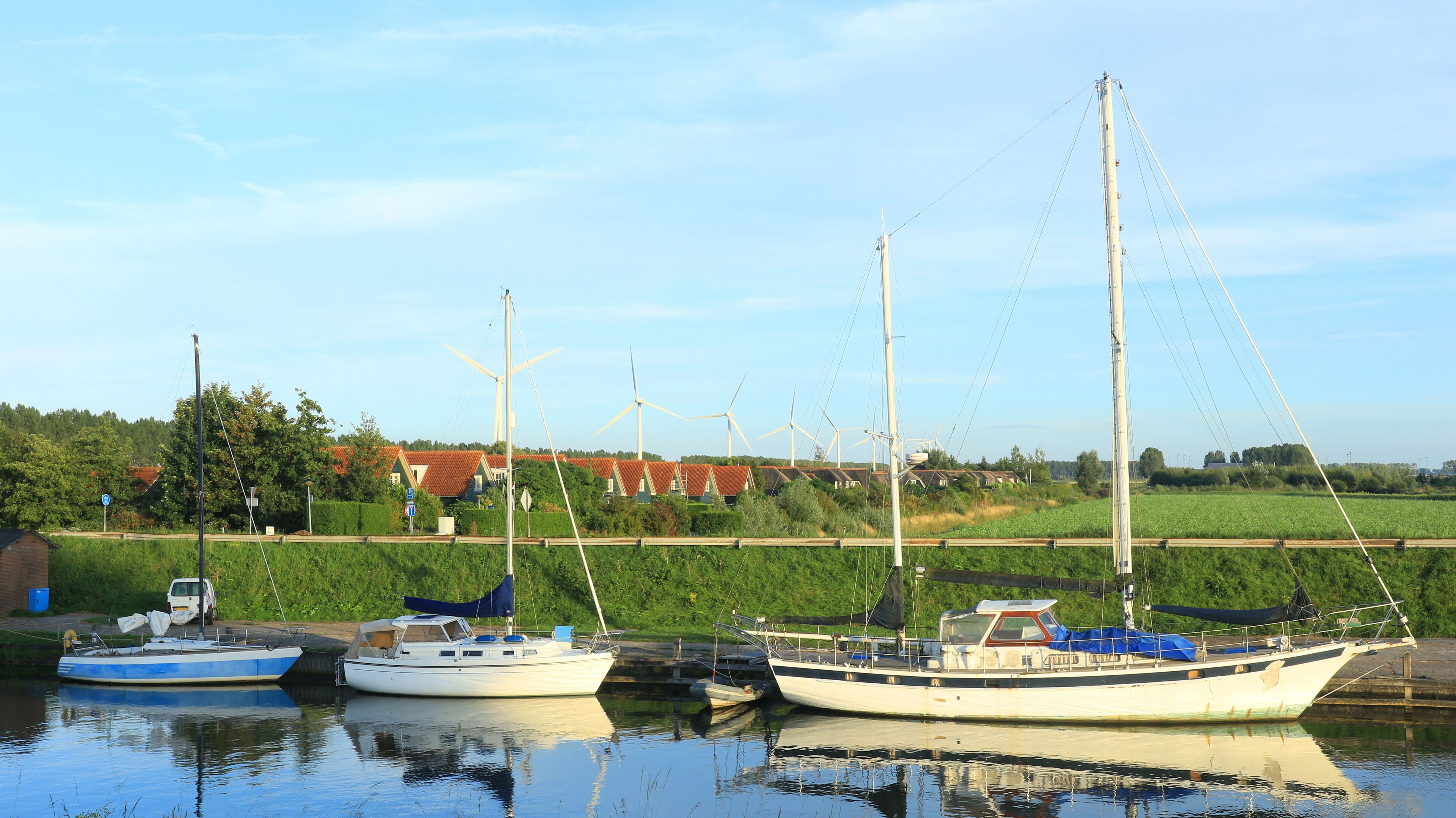 A calm harbor with two sailboats docked, surrounded by lush greenery and wind turbines in the background.