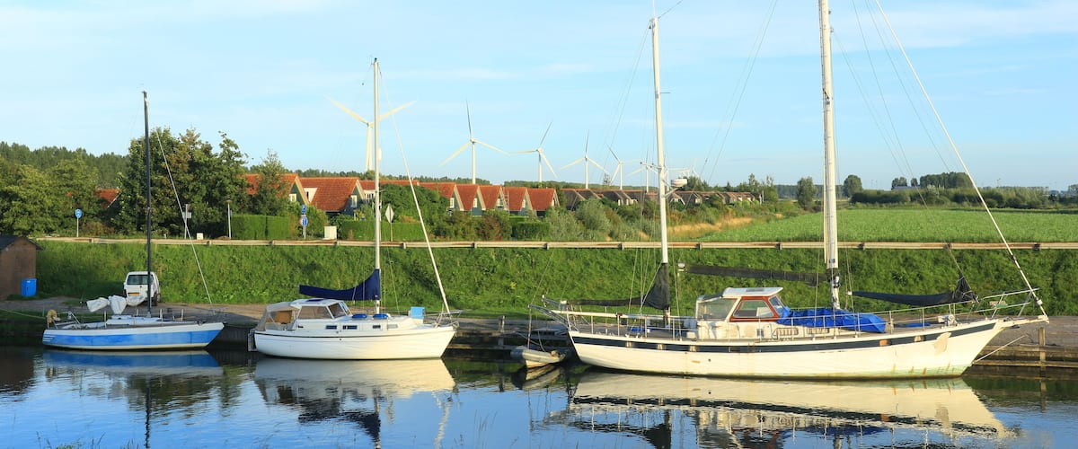 A calm harbor with two sailboats docked, surrounded by lush greenery and wind turbines in the background.