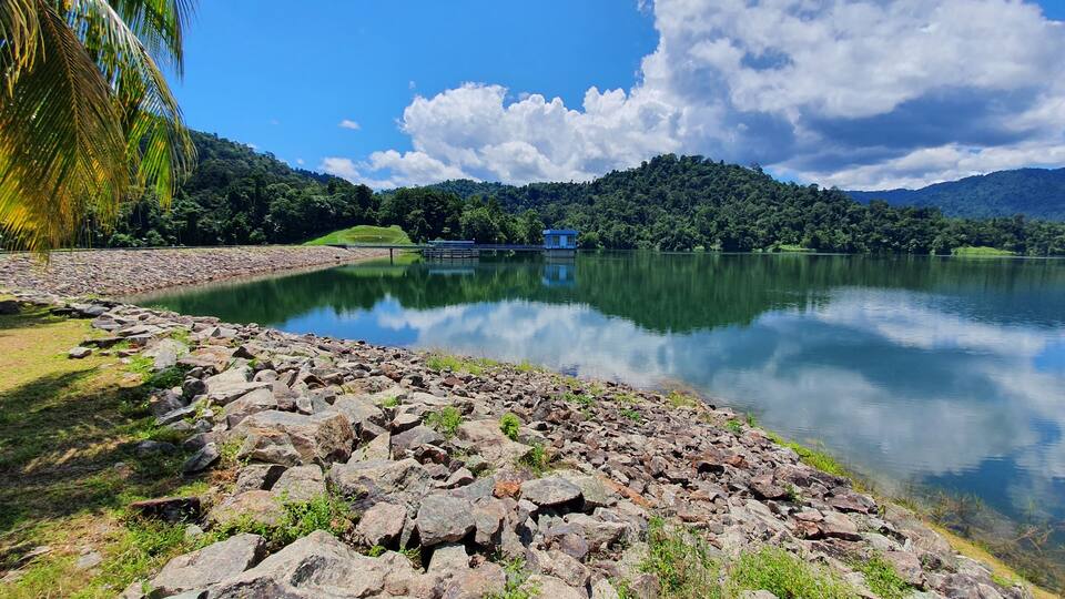 A calm lake with a rocky shoreline, a small blue house on the opposite shore, and lush green hills in the background.