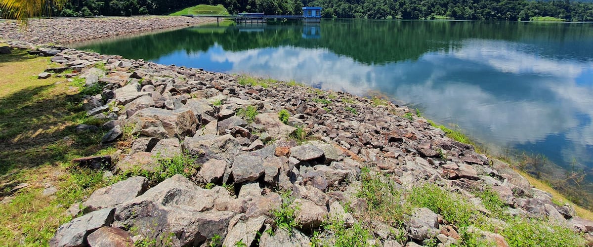 A calm lake with a rocky shoreline, a small blue house on the opposite shore, and lush green hills in the background.
