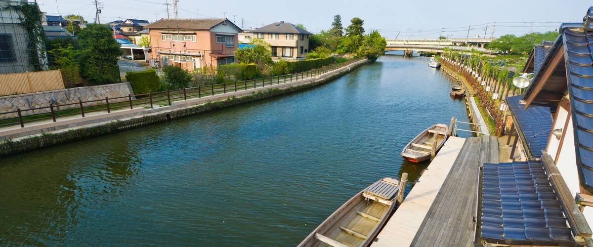 A calm river with boats docked along the shore, surrounded by residential buildings and a bridge in the background.