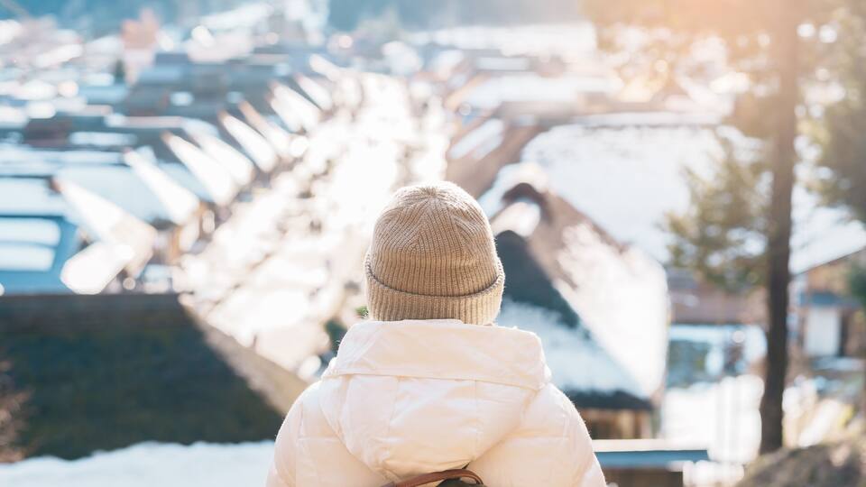 A person in a white jacket and beanie looking at a snow-covered landscape with houses.