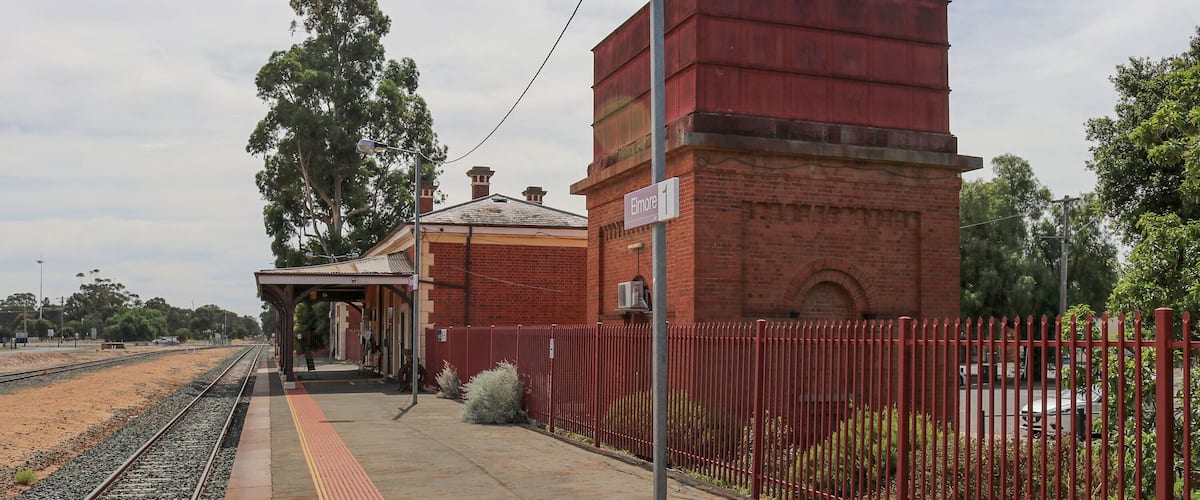 A railway station platform with a red brick building, a fence, and a large tree in the background.