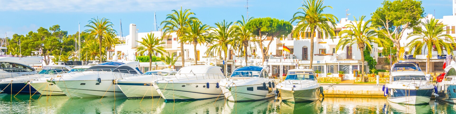 A marina with several yachts docked, surrounded by palm trees and white buildings under a clear blue sky.