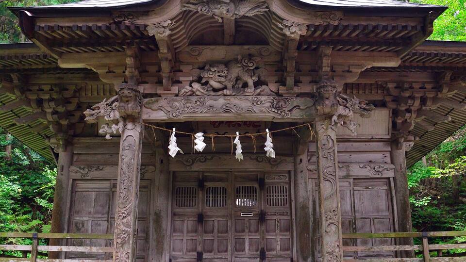 A traditional wooden shrine with ornate carvings and a tiled roof, surrounded by lush greenery.