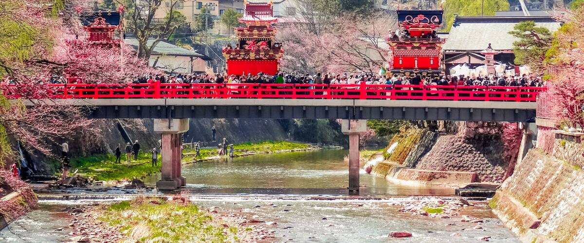 A bridge with traditional red structures, a river below, and cherry blossoms in the background.
