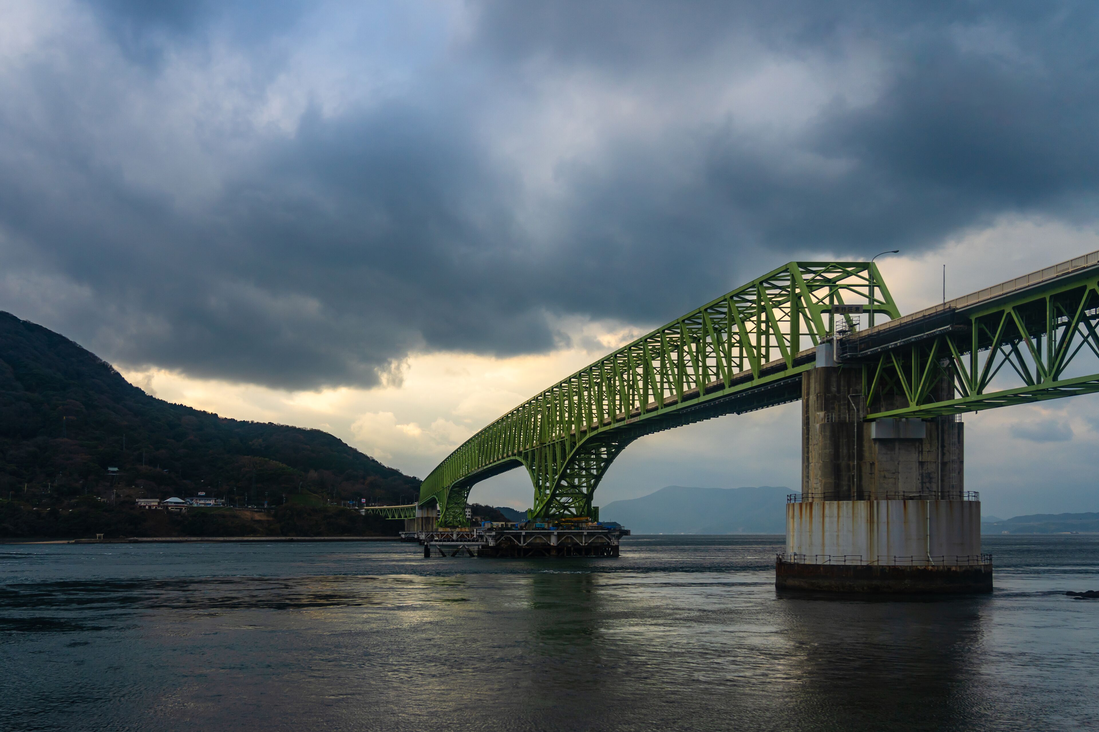 A green steel bridge spanning a body of water with mountains in the background.