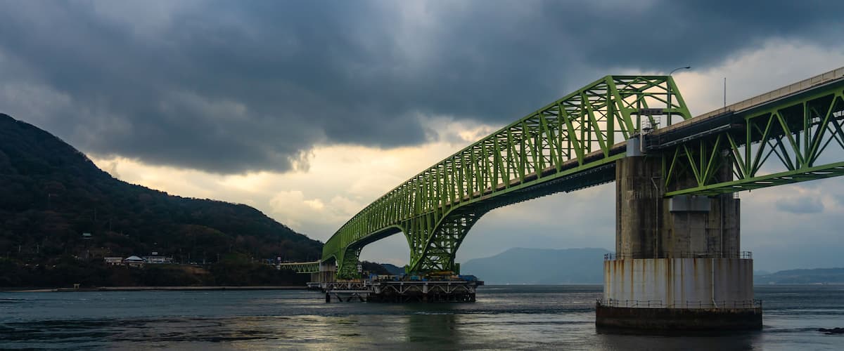 A green steel bridge spanning a body of water with mountains in the background.