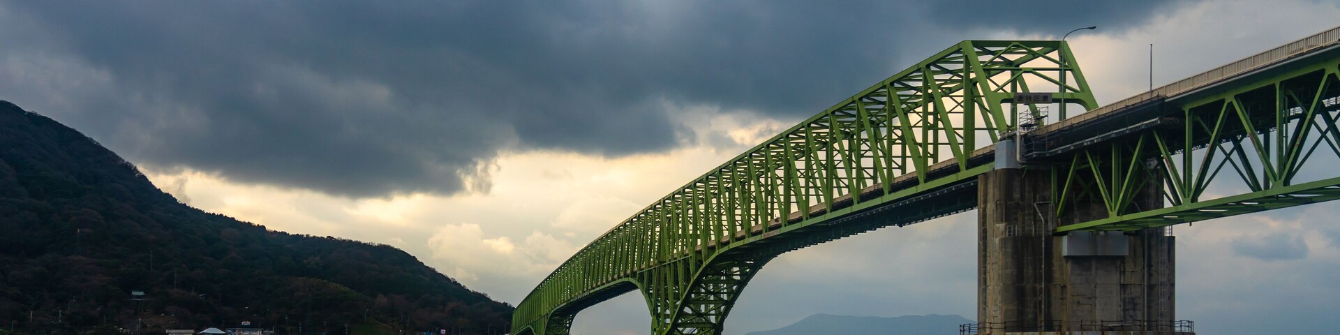 A green steel bridge spanning a body of water with mountains in the background.