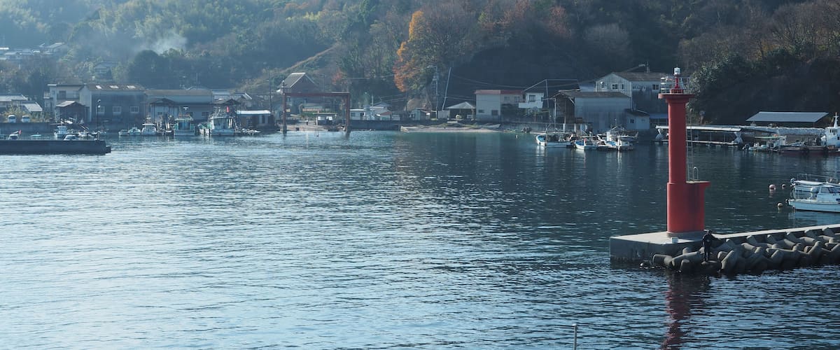 Un port côtier avec des bateaux, un phare et une chaîne de montagnes en arrière-plan.