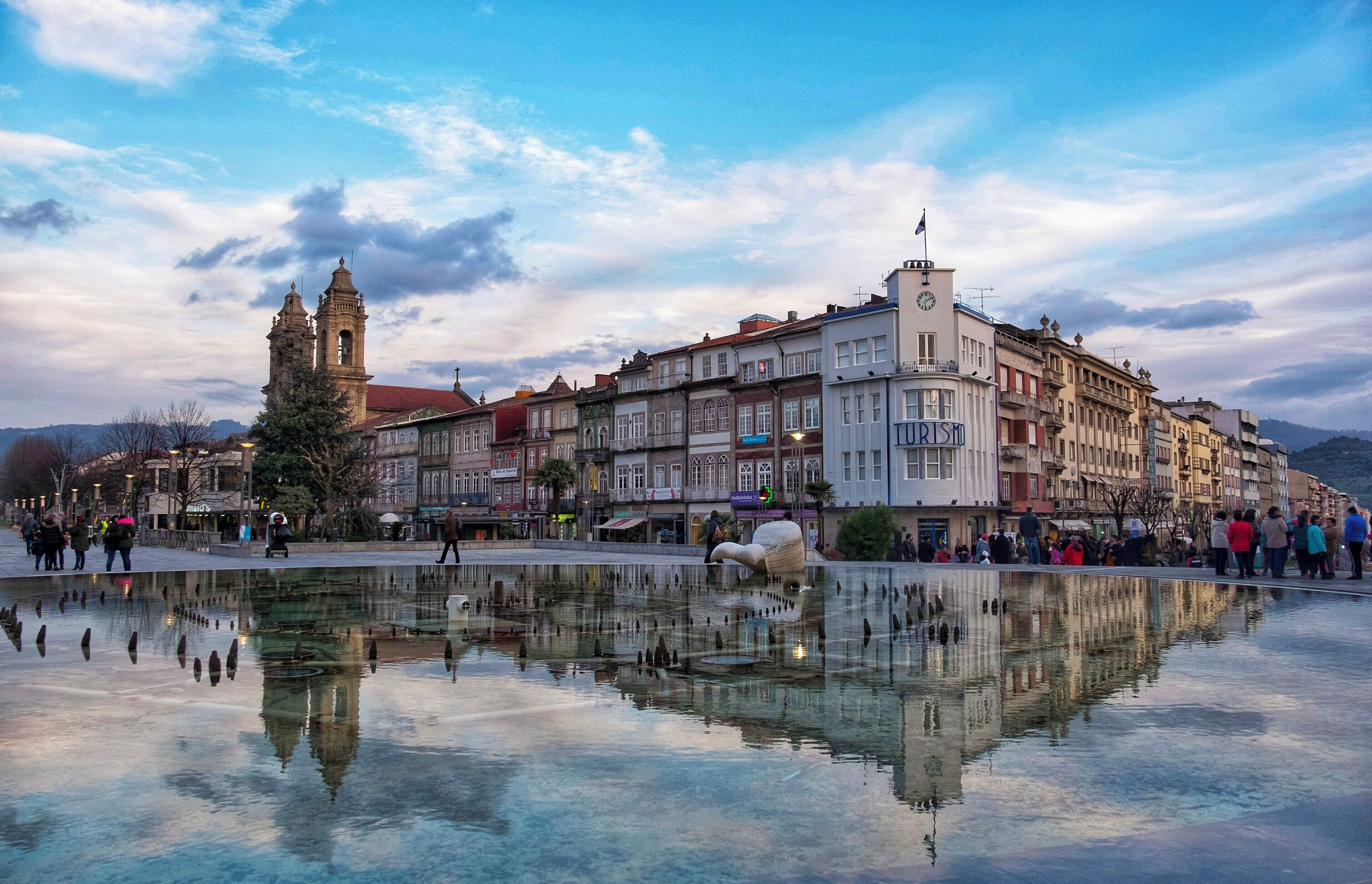 A historic town square with a central fountain reflecting buildings and a prominent clock tower.