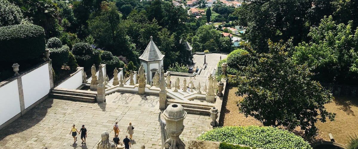 A historic garden with ornate statues, a central fountain, and a panoramic view of a city and mountains in the background.