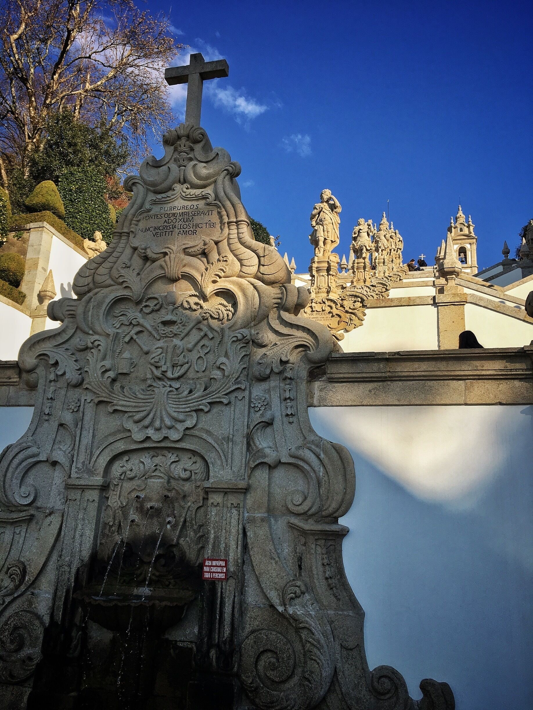 An ornate stone structure with a cross, intricate carvings, and a sign in a foreign language.