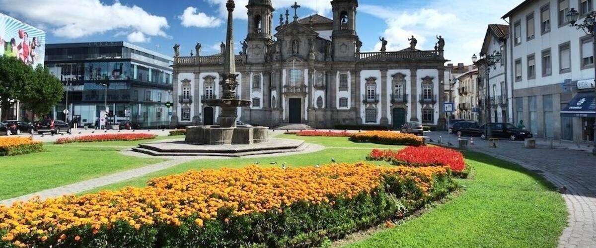 A historic church with twin towers and a central fountain surrounded by flower beds.