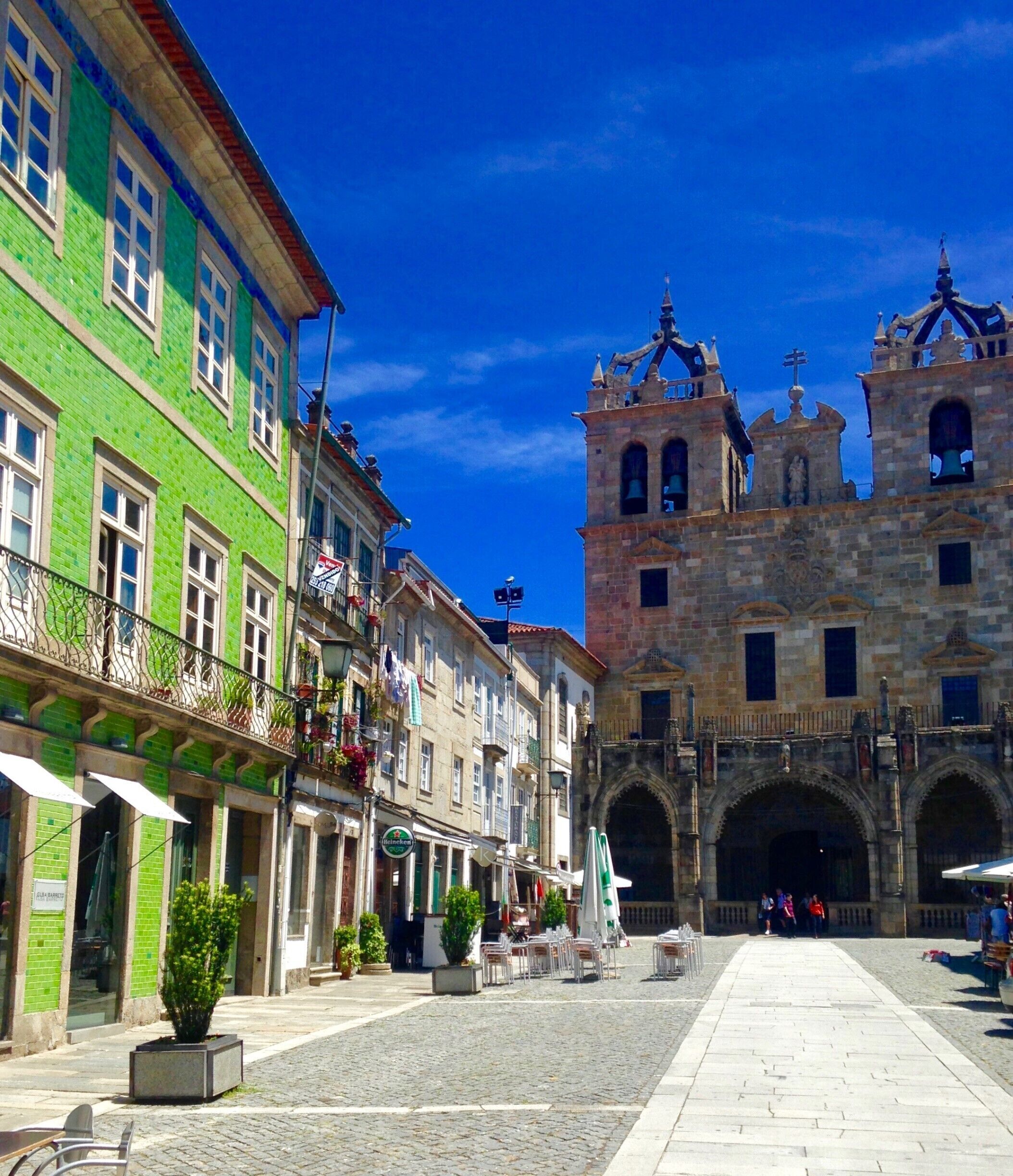 Uma praça de pedra com edifícios históricos, uma torre de igreja imponente e áreas de estar ao ar livre.