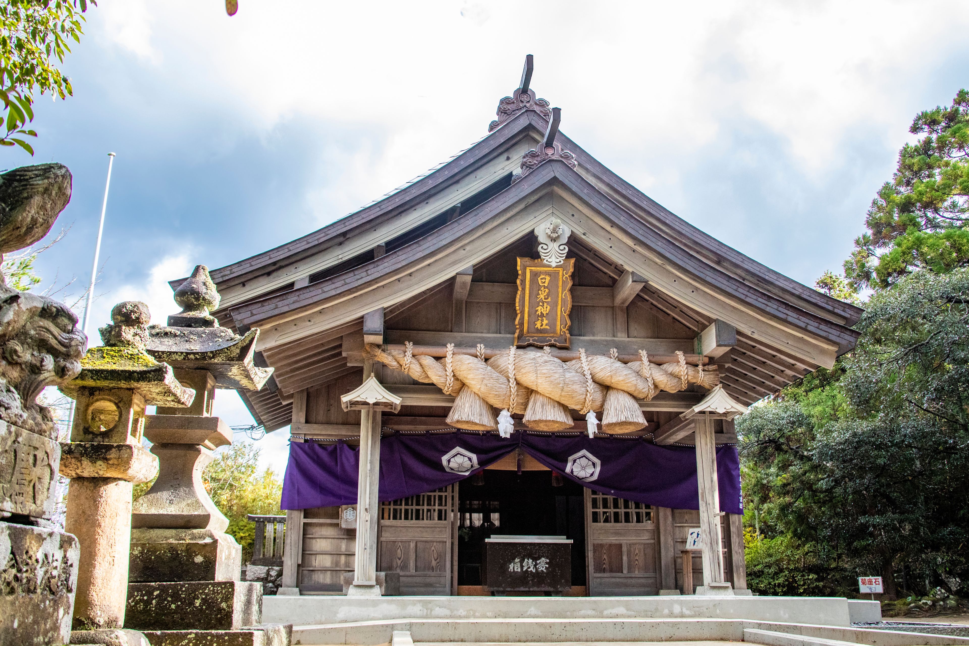 Perinteinen japanilainen temppeli, jossa on puinen rakennelma, japaninkielisiä merkkejä sisältävä kyltti ja violetti markiisi.