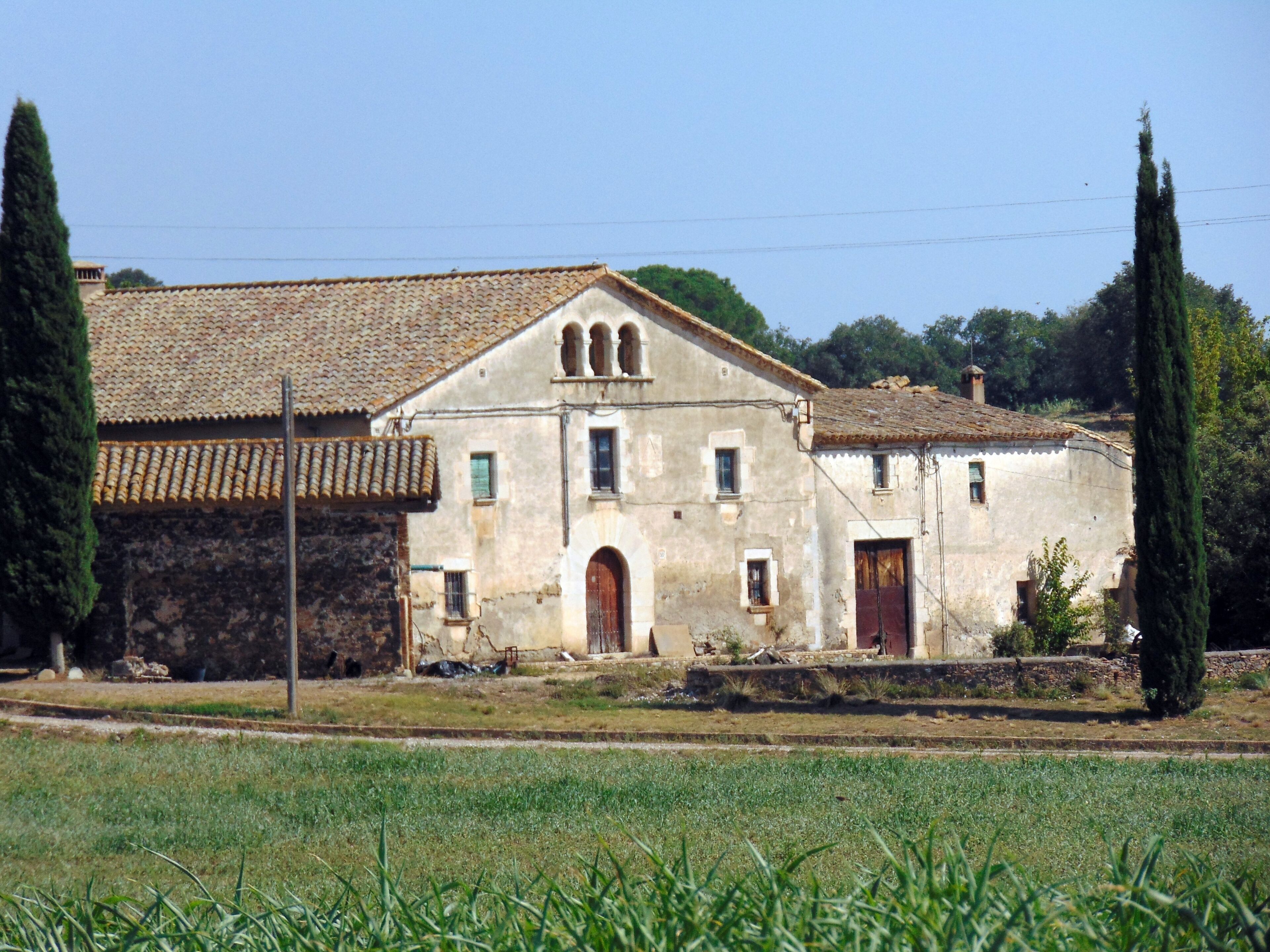 A rustic building with a tiled roof and arched windows, surrounded by a grassy field and tall cypress trees.