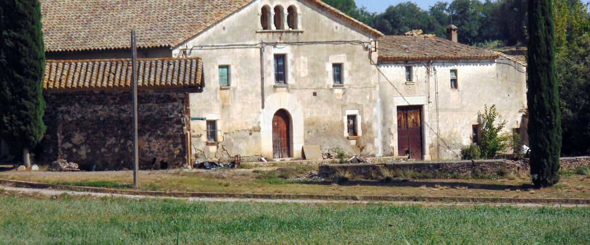A rustic building with a tiled roof and arched windows, surrounded by a grassy field and tall cypress trees.