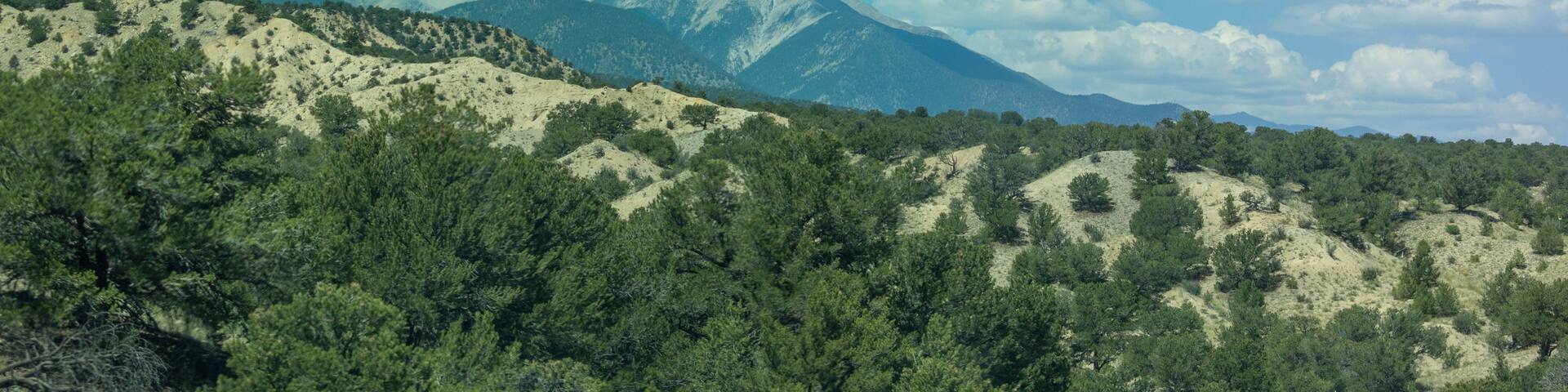 A mountainous landscape with dense green forests and a clear blue sky with fluffy white clouds.
