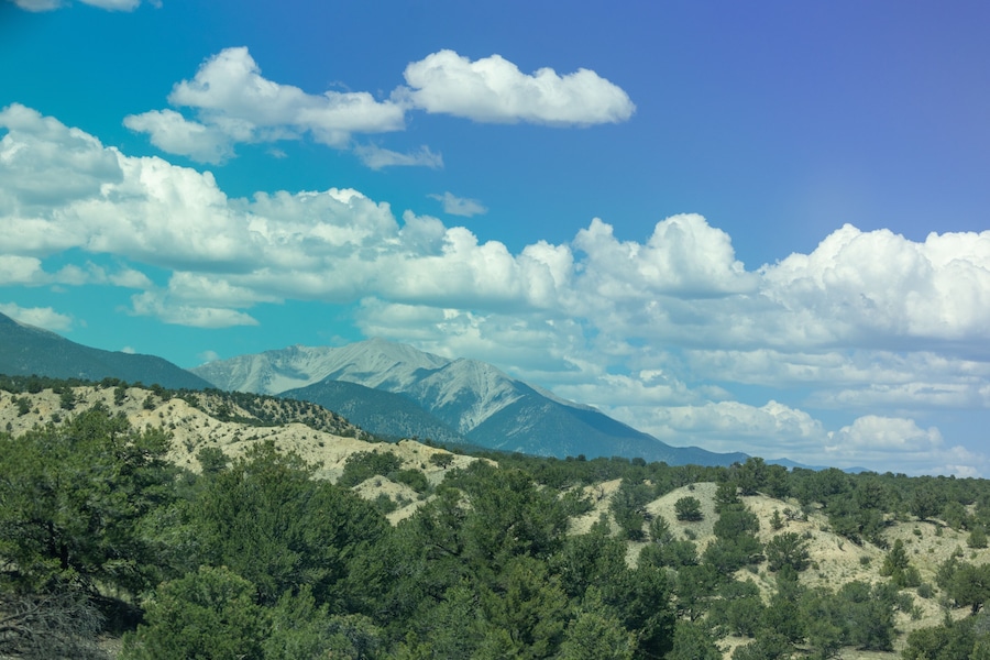 A mountainous landscape with dense green forests and a clear blue sky with fluffy white clouds.