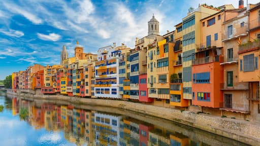 Un río refleja edificios de colores, una torre de iglesia y un cielo despejado.