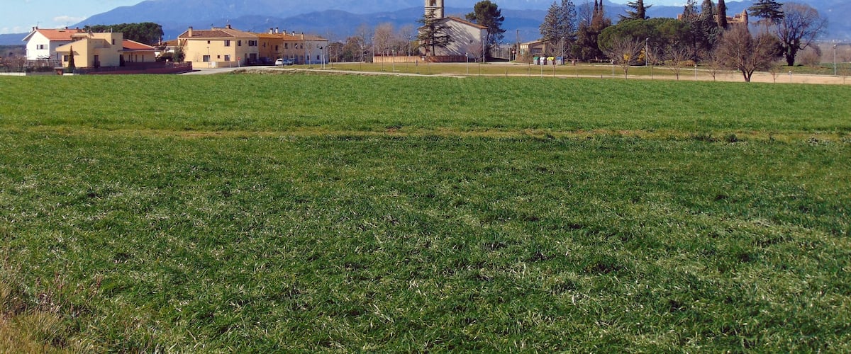 A rural landscape with a church, buildings, and snow-capped mountains in the background.