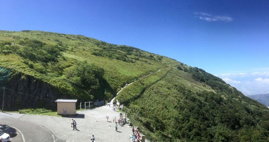 Un paisaje montañoso con una carretera pavimentada, un edificio pequeño y un grupo de personas caminando por un sendero.