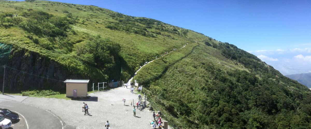 A mountainous landscape with a paved road, a small building, and a group of people walking along a trail.