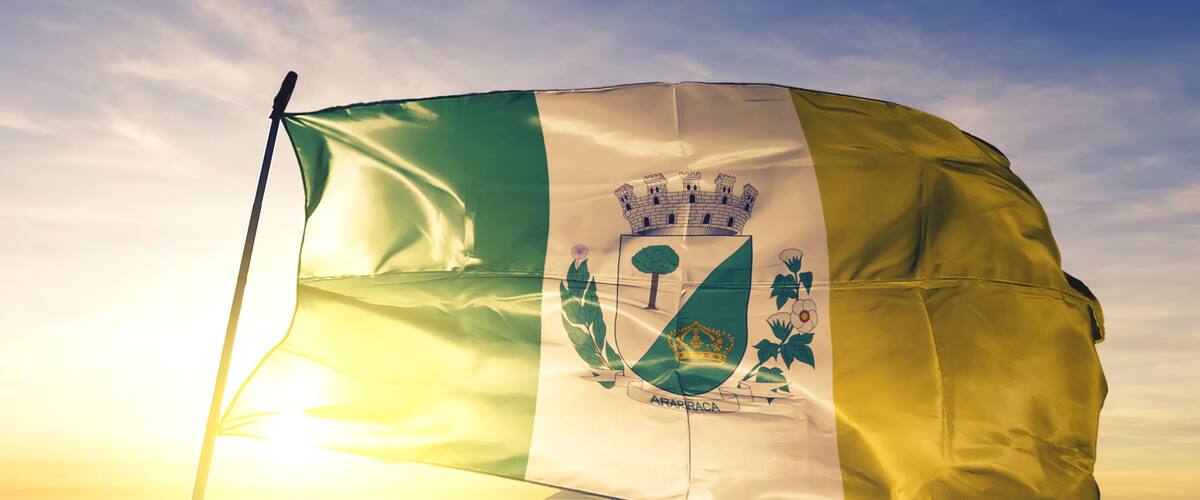 A flag with a crown, a shield, and the word "ARAPIRACA" on a pole against a sunrise sky.
