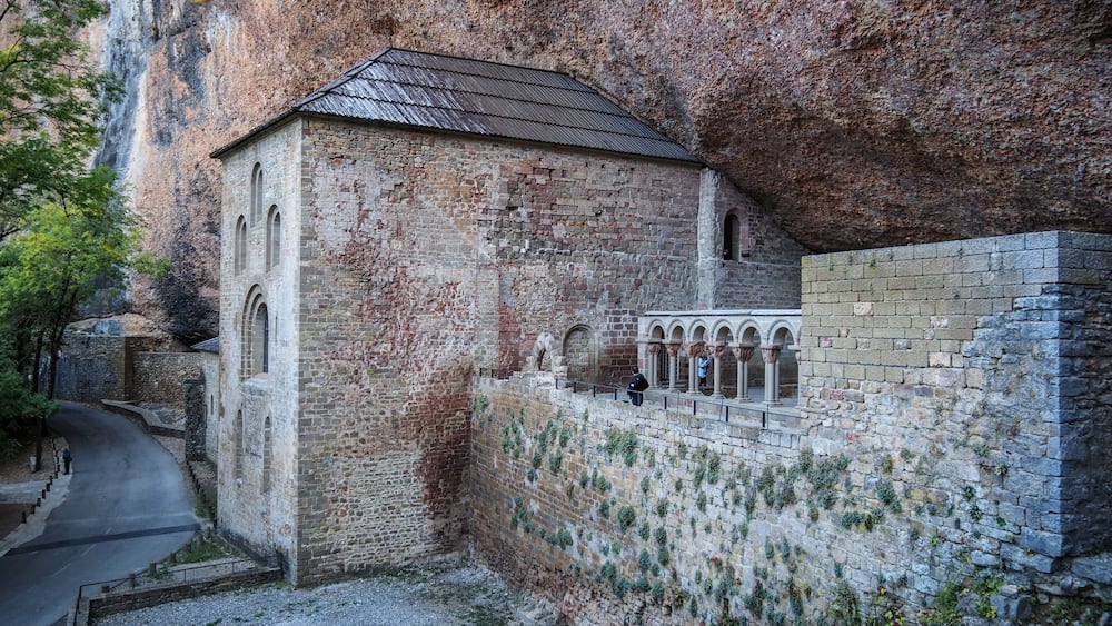 Ein historisches Steinhaus mit Ziegeldach neben einer Steinmauer mit Rundbögen.
