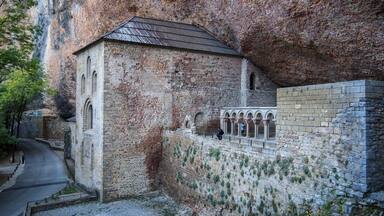 A historic stone building with a tiled roof, adjacent to a stone wall with arched niches.