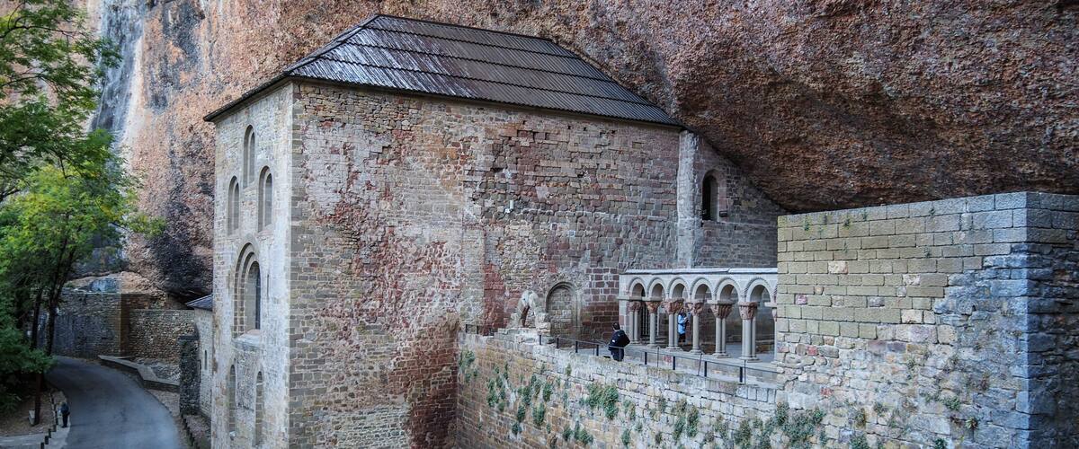 A historic stone building with a tiled roof, adjacent to a stone wall with arched niches.