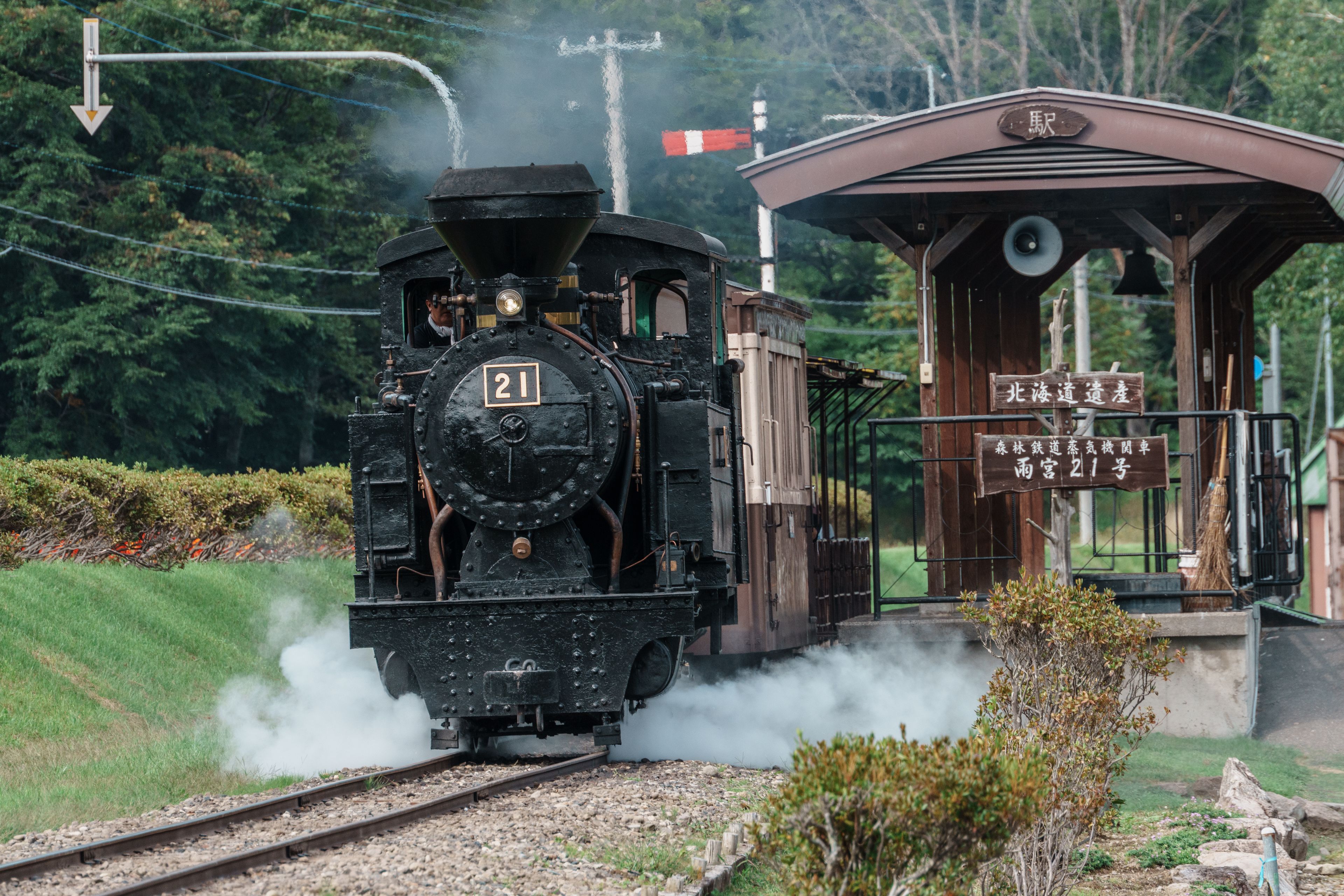 A steam locomotive numbered 21 moving on tracks with steam emission, passing by a station with a sign in a foreign language.