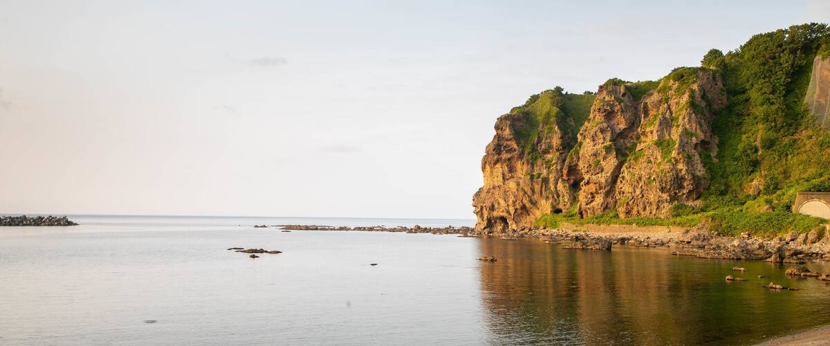 A coastal landscape with a rocky cliff, calm sea, and a sandy beach where people are walking.