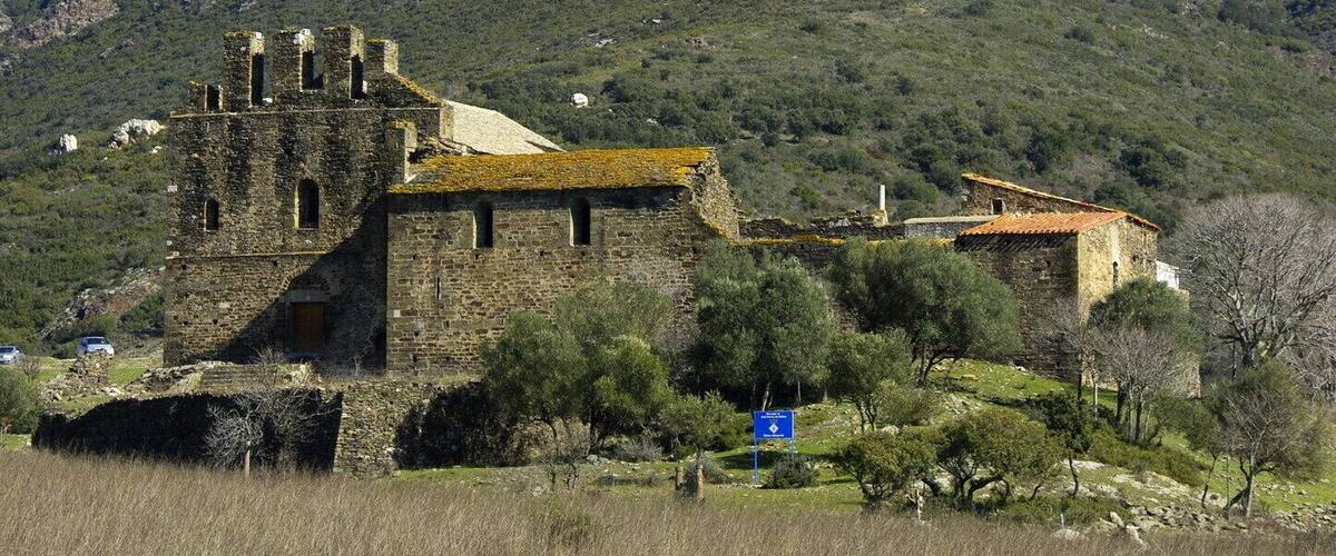 A historic stone building with ruins, surrounded by a rural landscape and a mountain in the background.