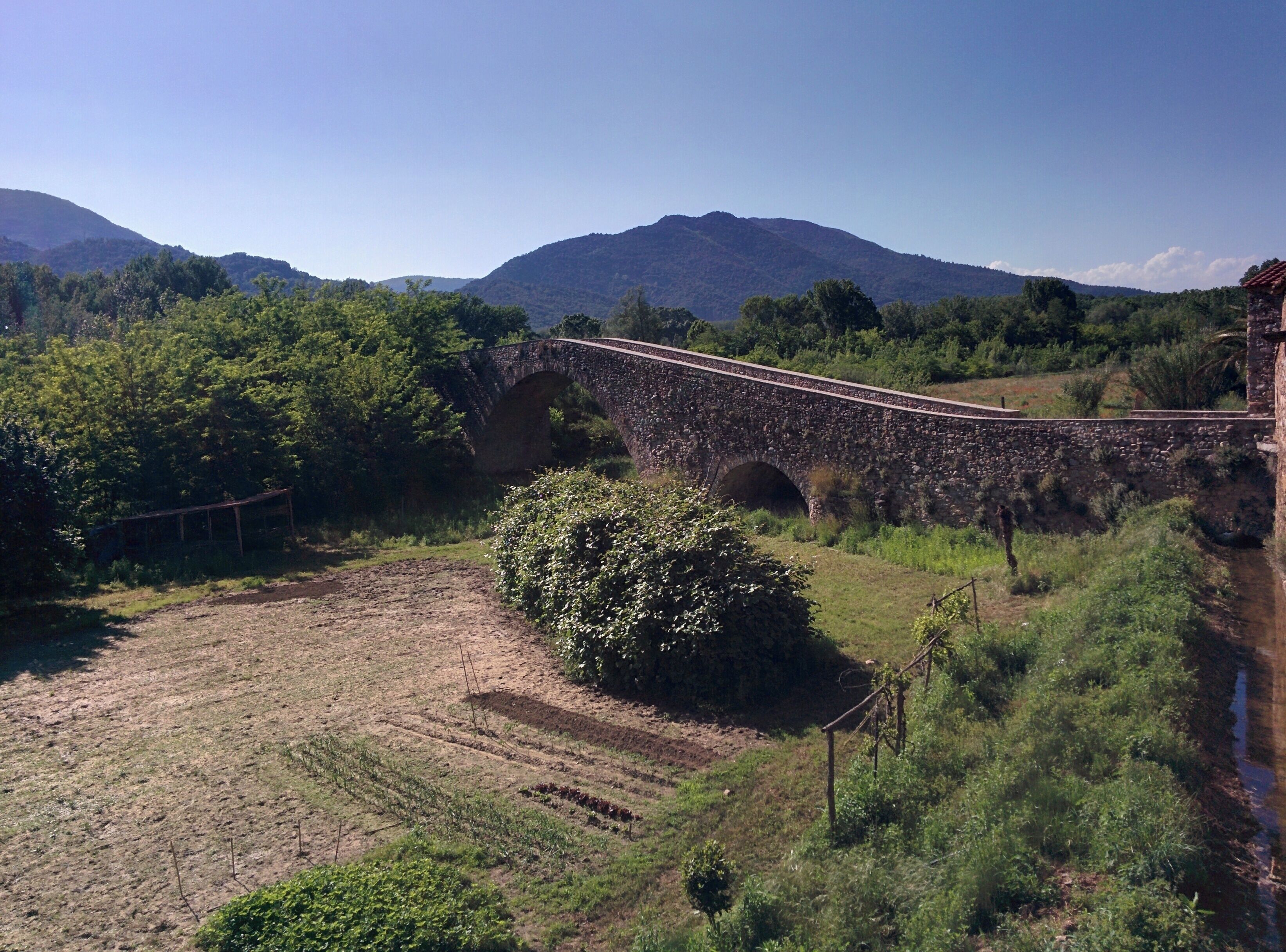 A stone bridge over a river, surrounded by lush greenery and mountains in the background.
