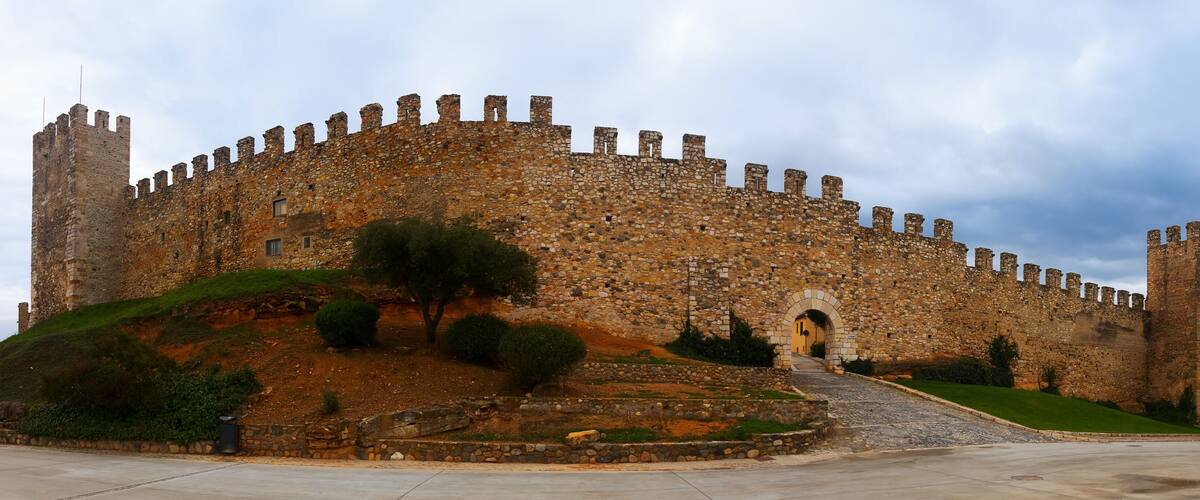 A historic stone castle with a large entrance gate, surrounded by a well-maintained garden and a paved pathway leading to the gate.