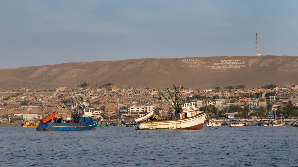 A coastal town with fishing boats, a prominent hill with inscriptions, and a communication tower in the background.