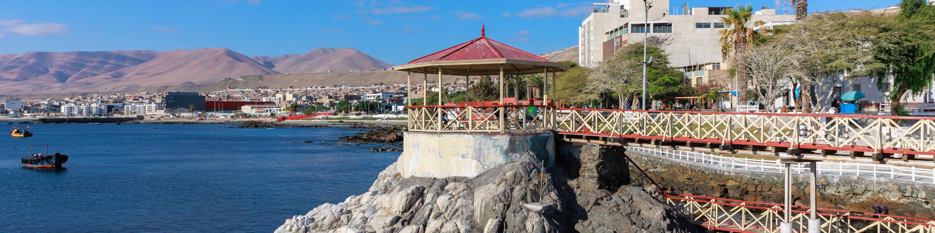 Zona costera con una formación rocosa, un pabellón de techo rojo, un puente y un paisaje urbano al fondo.